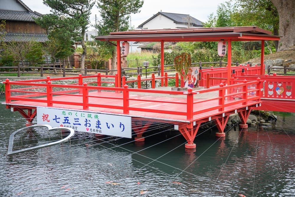 日本九州-戀木神社｜紅色的水上涼亭、木棧橋、橫幅標語、燈籠、水池、錦鯉、綠樹、日式建築。陰天戶外光線，紅色結構與灰色水面