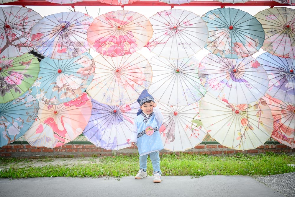 花蓮張家韓園｜街道、紙傘、孩童、傳統服飾，一名身穿淺藍色古裝的幼童站在整面懸掛色彩繽紛油紙傘的牆面前，低頭擦拭眼角