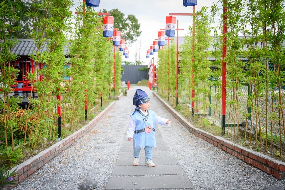 花蓮張家韓園｜小孩、步道、竹林、燈籠。身穿藍色傳統服飾的小孩站在碎石步道中央，兩側為綠竹與紅柱懸掛彩色燈籠。