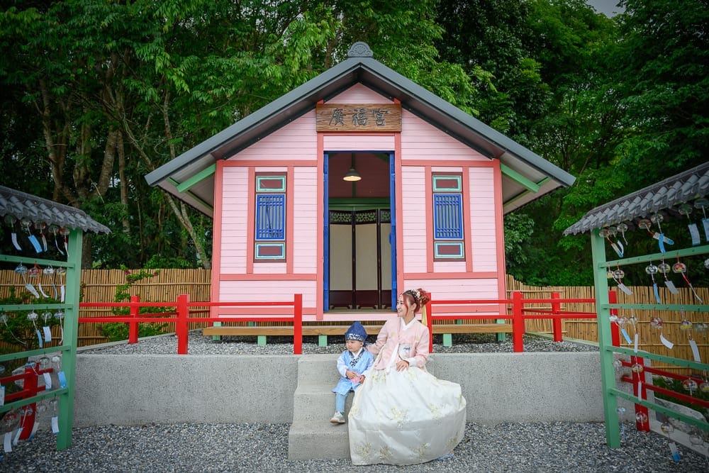 花蓮張家韓園｜女性與小孩身著傳統服飾，坐在粉紅牆面建築的石階前。建築屋簷懸掛木製牌匾，兩側架設懸掛祈福卡片的木架，
