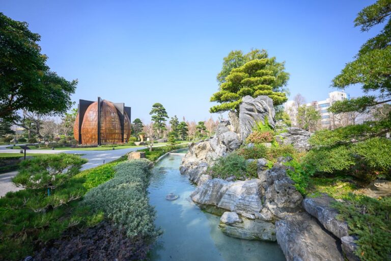 彰化成美文化園｜戶外庭園，小溪流、奇石堆疊、修剪整齊的松樹、大型竹編圓顶裝置藝術、草皮步道，晴朗藍天，遠處可見白色建