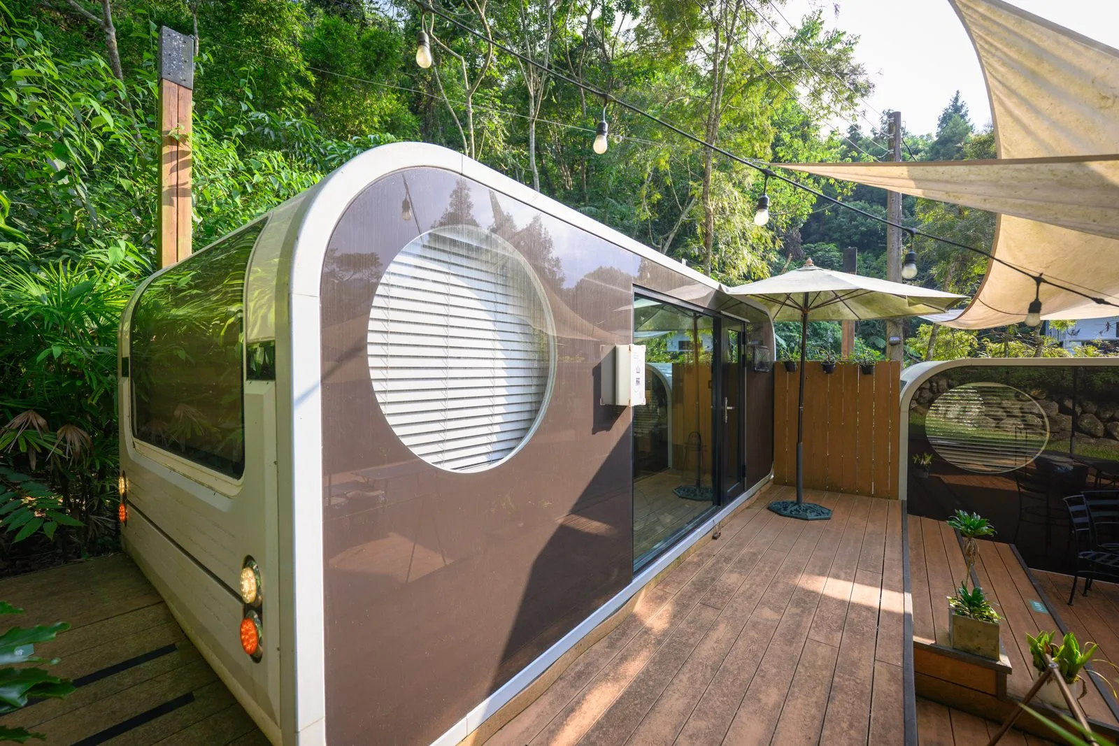 台中薰衣草森林住宿及園區｜香草House 露營車房型外觀、木棧平台、米色遮陽帆布、圓形車窗、串燈、遮陽傘。銀綠色露營車停在木棧平台上，車身開有大面圓窗。