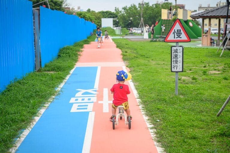 苗栗後龍(親子)六號公園完整指南|平衡車跑道與兒童遊樂設施玩法解析 10 苗栗後龍六號公園|兒童平衡車、跑道、藍色圍籬、草地、警告標誌、涼亭,戶外公園,一名幼童騎乘平衡車行駛於紅藍相間的專用道