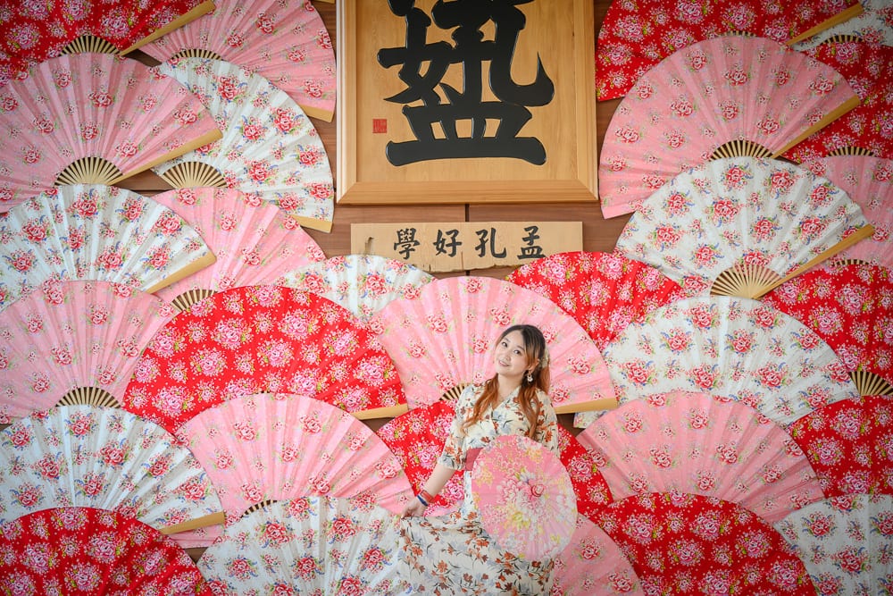 彰化成美文化園｜女子、扇子、木匾、牆面，女子身穿印花洋裝，手持同款粉紅圓傘，置身於佈滿紅粉花卉摺扇的牆前，正中央上方