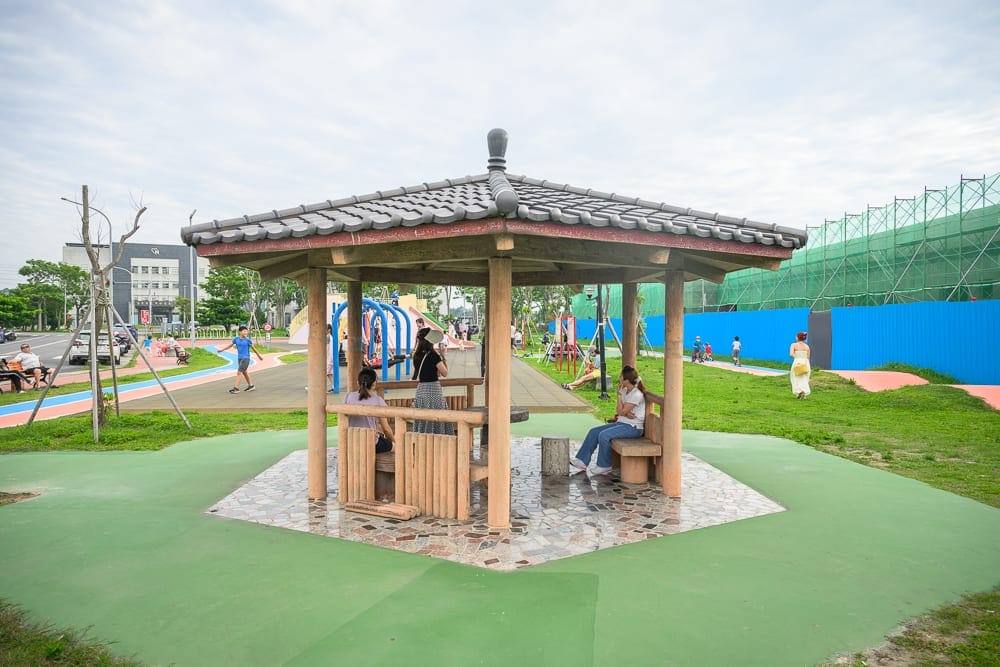 苗栗後龍(親子)六號公園完整指南|平衡車跑道與兒童遊樂設施玩法解析 6 苗栗後龍六號公園|戶外公園、涼亭、座椅、草皮、行人、溜滑梯、藍天。六角形木造涼亭坐落於綠色草皮上,周圍設有石椅,幾位民