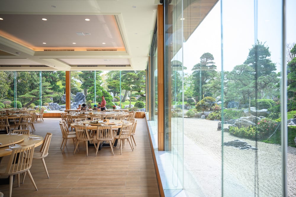 彰化成美文化園｜室內、餐廳、圓桌、木椅、落地窗、庭園、松樹、石塊，寬敞木質地板延伸至整面玻璃窗，窗外映出日式造景與綠