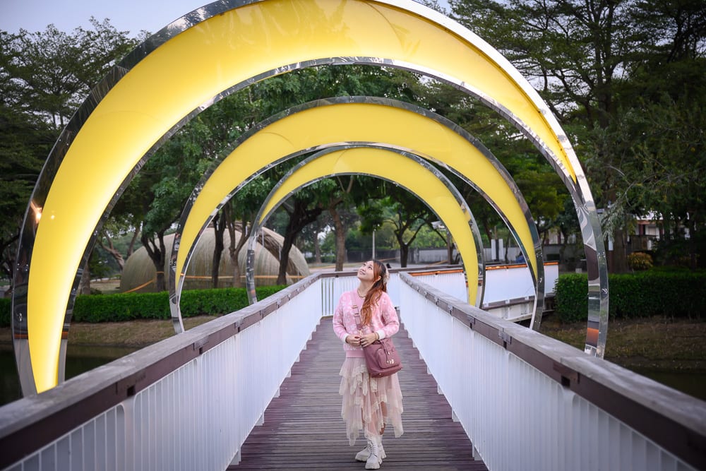 台南月之美術館｜女性、橋、黃色圓弧藝術裝置、綠樹、水面、公園。一名粉衣女子站在木棧道橋樑中央，抬頭望向頭頂上方層層排