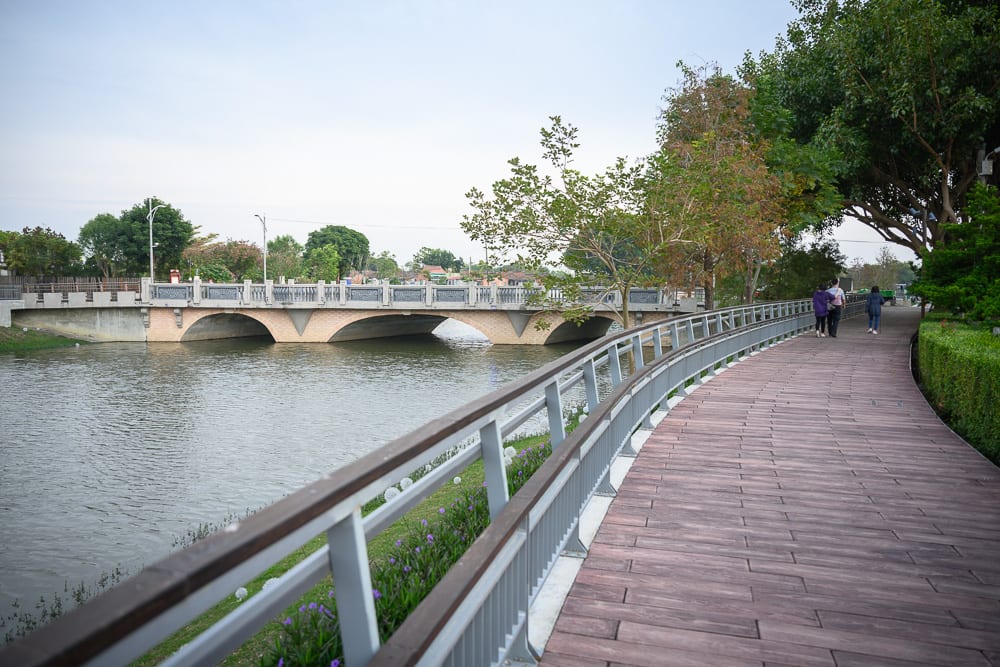 台南月之美術館｜木棧道、河、橋、欄杆、行人、樹木，沿著河岸延伸的木造步道設有金屬扶手，幾位行人正向前漫步。前方橫跨著