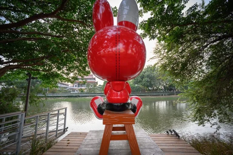 台南月之美術館｜大型紅色兔子雕塑、木板凳、河畔、木棧道、綠樹、欄杆、建築物，兔子背對鏡頭坐在河邊木棧道的高腳凳上，上