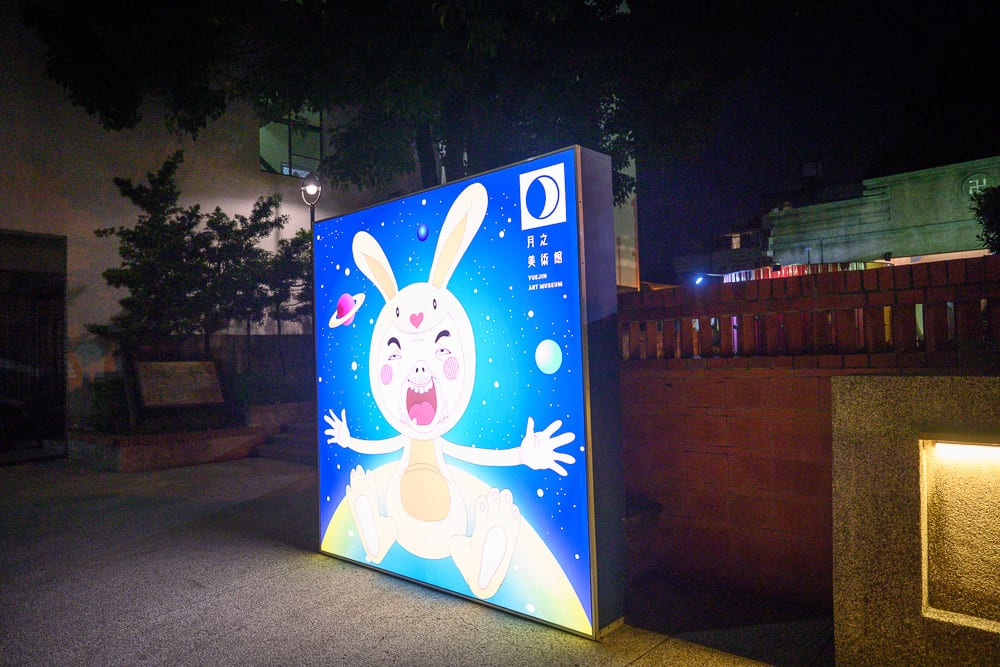 台南月之美術館｜夜晚的街道、燈箱、兔子角色、磚牆、樹木，亮著藍光的巨大矩形招牌立於路旁，畫面中央印有誇張表情的兔耳角