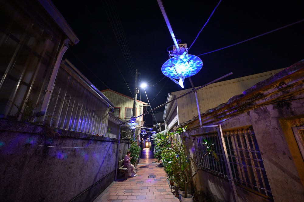 台南月之美術館｜夜間狹窄街道，磚造平房牆壁，電線杆，懸掛藍色發光圓形投影裝置，路燈，盆栽植物，坐於長椅上的女子。