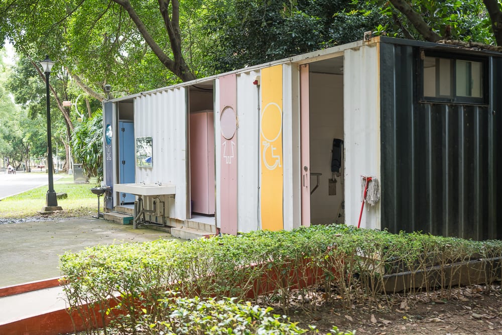 新北林口樂活公園｜貨櫃屋改造的公共廁所，設置於綠意盎然的公園步道旁，搭配矮灌木叢、路燈、洗手台，外牆漆上黑白與粉黃色調