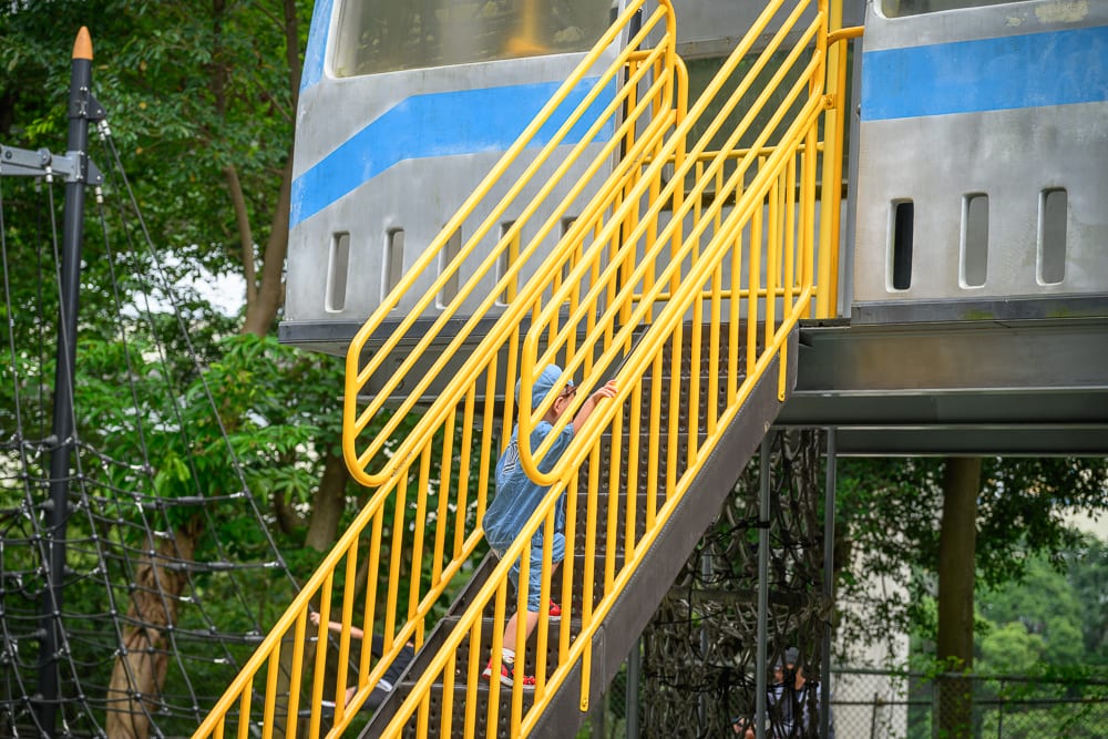 新北林口樂活公園｜火車造型遊具、金屬樓梯、繩網、綠樹、公園，兒童攀爬紅色鞋子階梯，欄杆排列平行線條，周圍林木茂密陰影錯