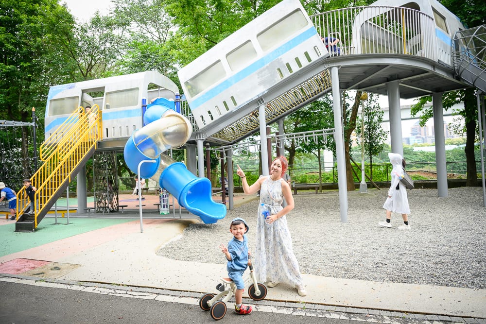 新北林口樂活公園｜婦人、男童、三輪車、溜滑梯、捷運車廂遊具、公園、樹木。穿著長裙的婦人與騎三輪車的男童站在大型捷運車廂