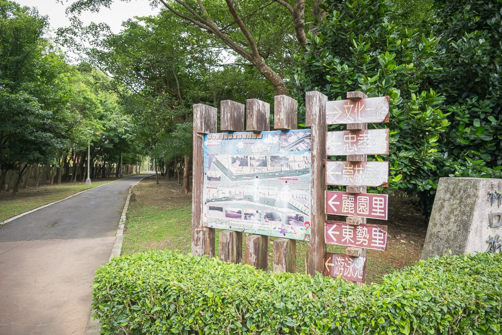 新北林口樂活公園｜木製指示牌、地圖告示牌、柏油小徑、綠色灌木叢、茂密樹林，戶外公園旁立著多向指標與全區導覽圖，左側道路