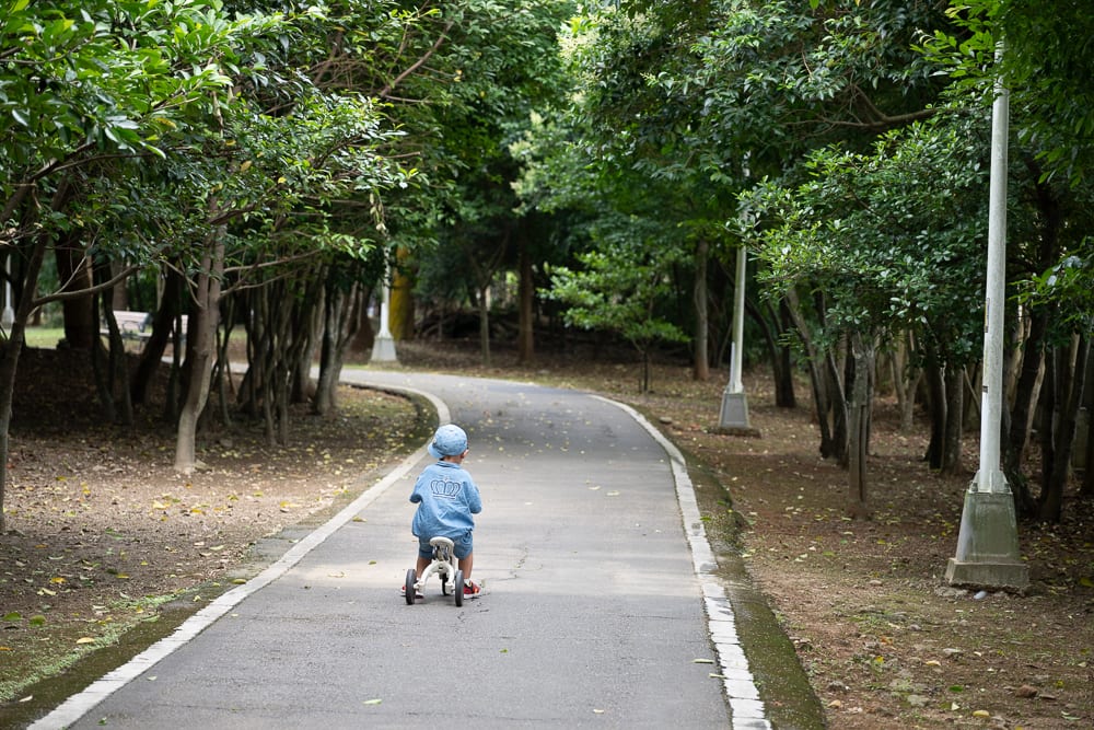 新北林口樂活公園｜小孩、平衡車、柏油路、樹林、路燈，戴藍帽與穿藍衣的幼童背對鏡頭，騎著無踏板的滑步車，行駛在兩側佈滿綠
