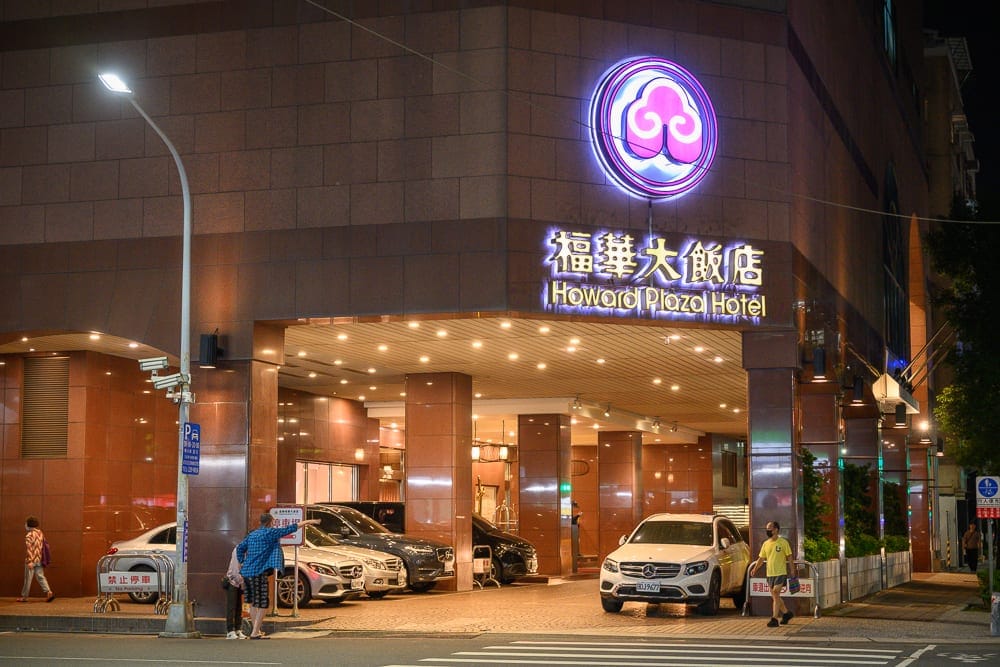 高雄福華大飯店完整攻略|屋頂泳池、早餐自助與尊爵房型解析 9 高雄福華飯店|街道,飯店,汽車,路燈,行人。夜間燈光照亮福華大飯店的褐色磁磚外牆與圓形招牌。騎樓下停放多輛轎車,戴