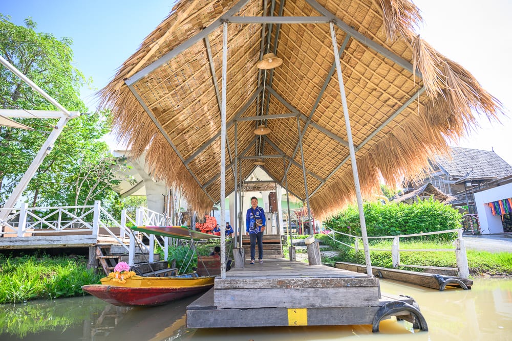 泰國芭達雅四方水上市場｜茅草屋頂、木棧道、河川、小船、男子，戶外。男子站在水中木棧道終點，上方搭設金屬支架與茅草遮陽棚，左側