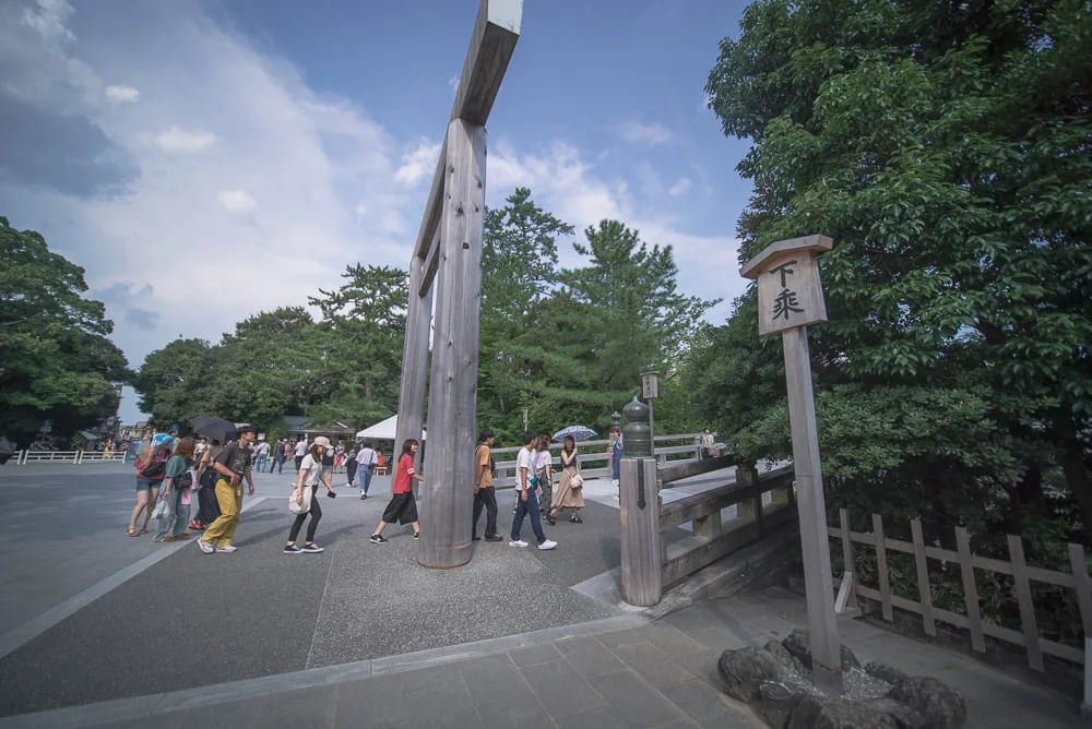 日本三重伊勢神宮｜戶外廣場，木造鳥居，下乘木牌，石板地，橋，人群，綠樹，藍天白雲。行人三三兩兩走過大型鳥居下方與橋梁入