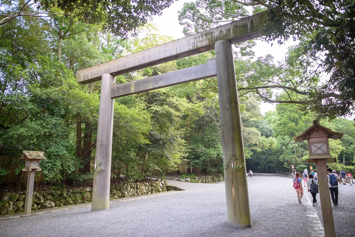 日本三重伊勢神宮｜神社、鳥居、林道、石燈籠、碎石路、遊客。高大的木造鳥居矗立於碎石路入口，兩側設置木製石燈籠。四周環繞