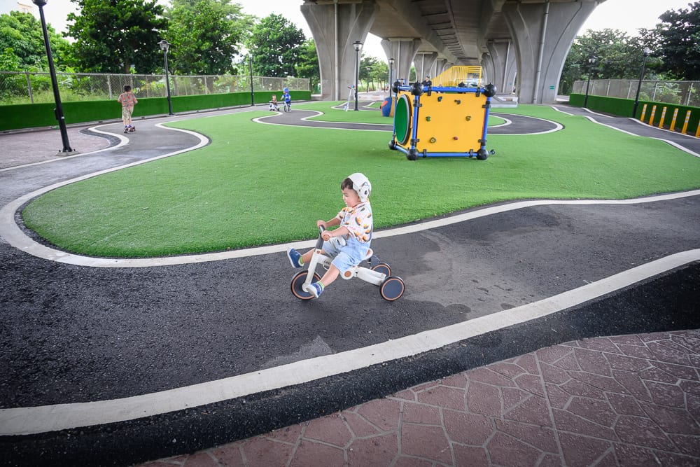 苗栗竹南滑步車公園｜橋下空間，幼童，三輪車，黑色跑道，人工草皮，黃色遊具。幼童戴安全帽騎乘白色三輪車行駛於弧形跑道，周圍