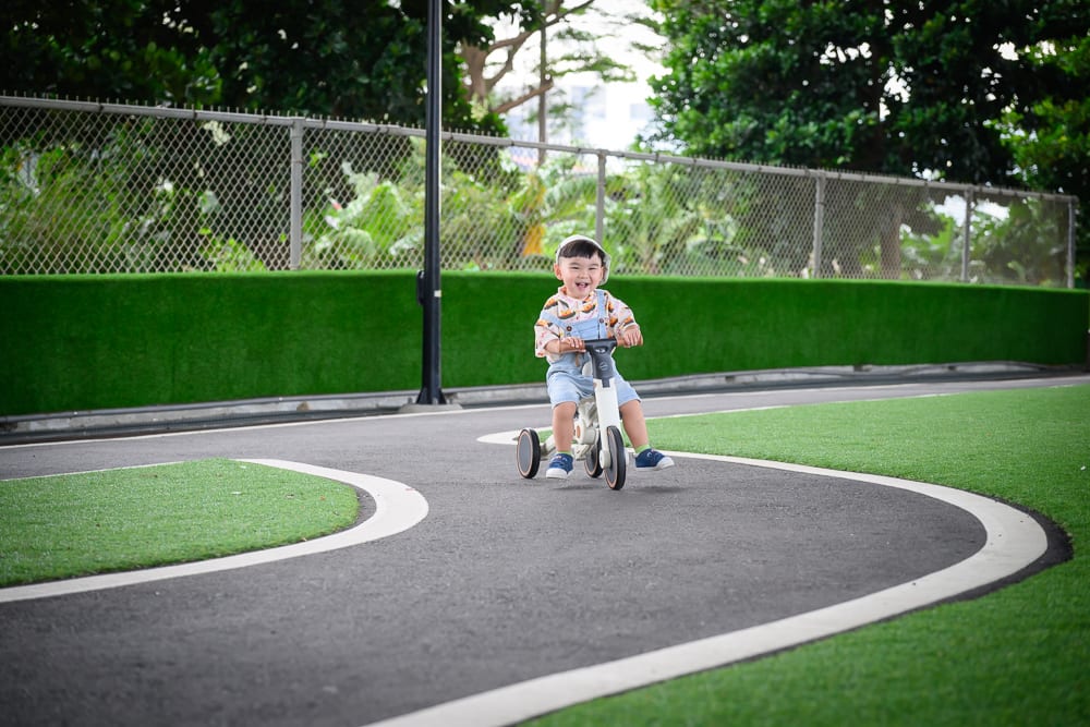 苗栗竹南滑步車公園｜兒童，三輪車，戶外賽道，人工草皮，鐵絲網圍籬，柏油路面，彎道。一名幼童騎著三輪車行駛在鋪有白色邊線的