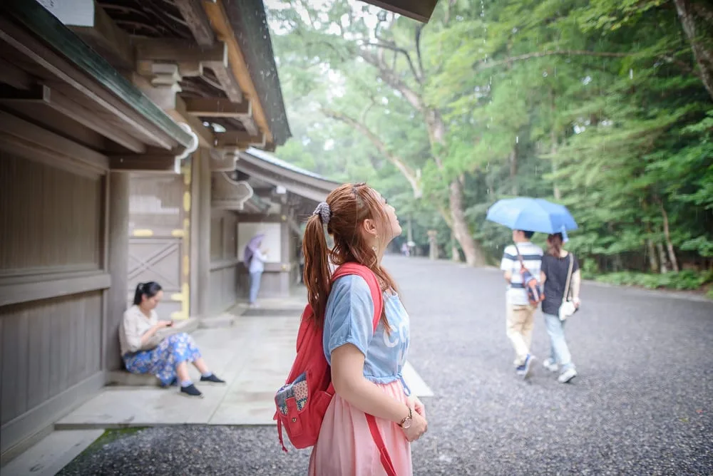 日本三重伊勢神宮｜木造走廊、紅背包女子、碎石路、藍雨傘、綠樹林、雨滴。扎馬尾的女子側身仰望，廊下坐著滑手機的人，遠處行