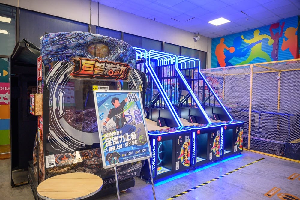 高雄 E7PLAY 三多館完整攻略|保齡球道、電子飛鏢與多元機台玩法解析 12 高雄E7play|室內,遊樂場,遊戲機台,投籃機,告示牌,網狀圍欄。亮藍色燈條發出藍紫光線,天花板嵌燈照射灰色水泥地坪