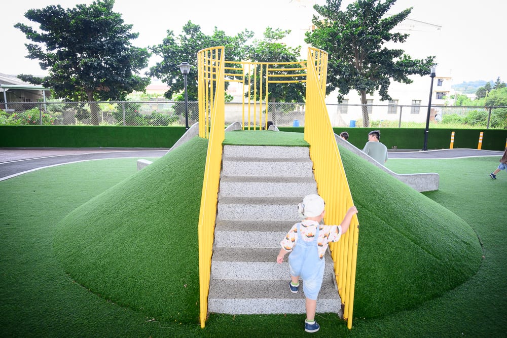 苗栗竹南滑步車公園｜兒童遊樂場，人工草皮，小丘，樓梯，溜滑梯，黃色欄杆。一名幼童正手扶黃色欄杆爬上鋪有綠色人工草皮的小丘