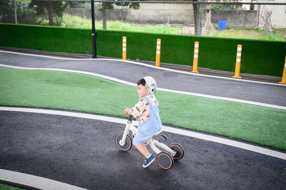 苗栗竹南滑步車公園｜小童，三輪車，柏油路，人工草皮，鐵絲網，黃白相間護欄。一名戴帽幼童穿著吊帶褲，騎乘白色三輪車行駛於鋪