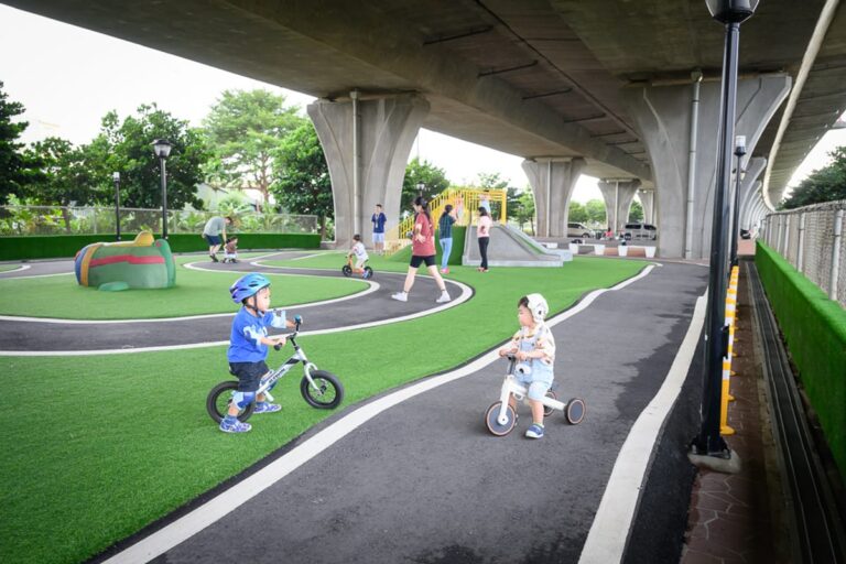 苗栗竹南滑步車公園｜高架橋下空間，人工草皮，滑步車道，幼童，家長，遊樂設施，路燈。兩名幼童騎著滑步車在黑色跑道上，四周鋪