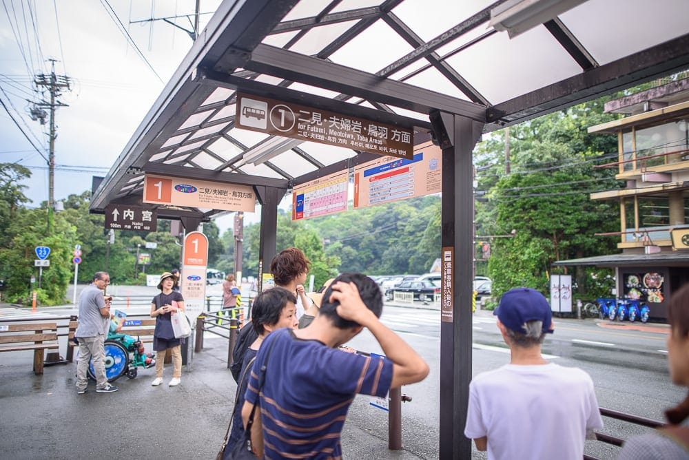 日本三重伊勢神宮｜公車站牌、候車亭、行人、指示牌、街道、建築。透明遮雨棚下有多位乘客排隊等車，頭頂懸掛棕色公車路線圖標