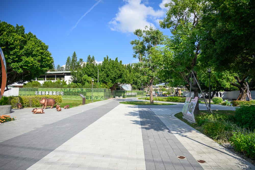 台中豬事圓滿公園｜戶外公園、石磚步道、綠樹、草皮、雕塑、藍天白雲。灰色與白色相間的石磚步道向前延伸，兩側綠樹成蔭並設有草皮，左側草地上散落著數隻咖啡色小豬雕塑，遠方可見低矮建築。