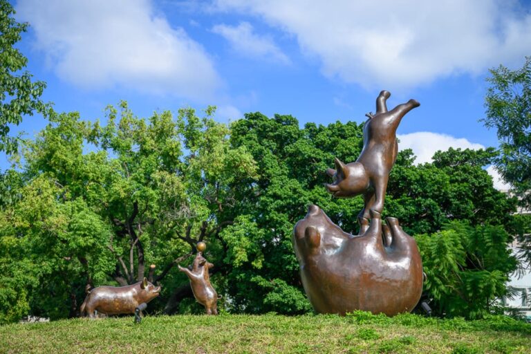台中豬事圓滿公園完整指南|共融遊戲場與銅豬雕塑 15 台中豬事圓滿公園|草地、雕塑、銅豬、樹林、藍天、白雲。綠色草皮上設置數座青銅豬隻雕塑,姿態各異,背景為茂密綠樹與湛藍晴空。