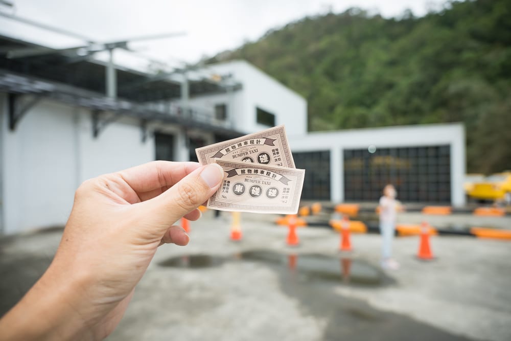 宜蘭計程車博物館｜手，票券，建築物，交通錐，戶外廣場。畫面前景為手部特寫，指間夾著兩張復古票券；背景為白色建築物、地面水漬與橘色交通錐，遠處可見綠意山景與模糊人影。