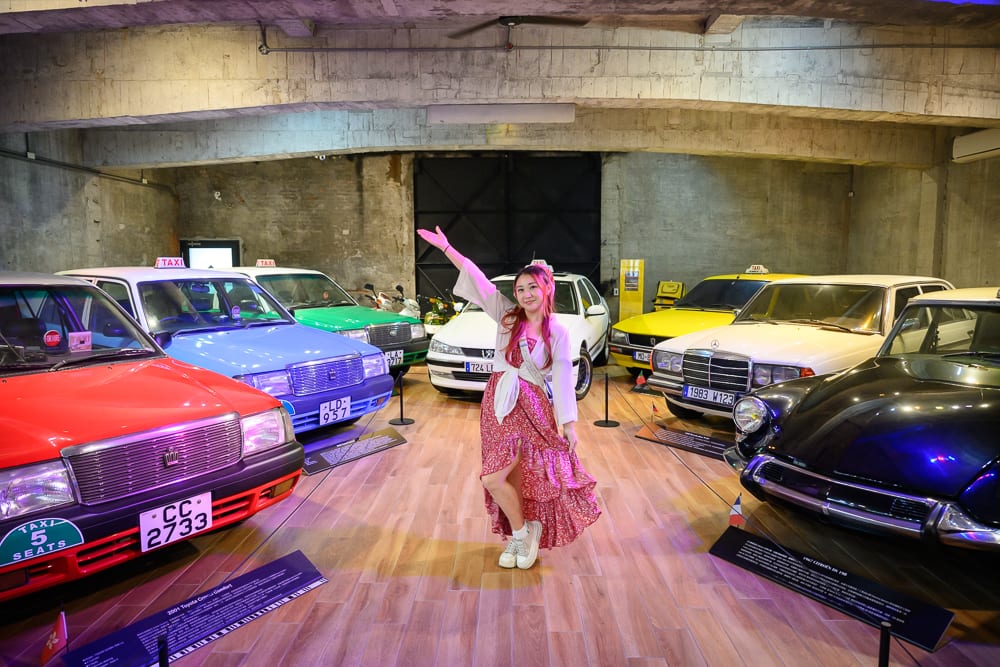 宜蘭計程車博物館｜室內展場、女子、古董車、計程車、木地板、水泥牆，女子站在多輛展示的彩色老舊計程車中間，一手高舉微笑，周圍環繞紅、藍、白、黑等車輛。