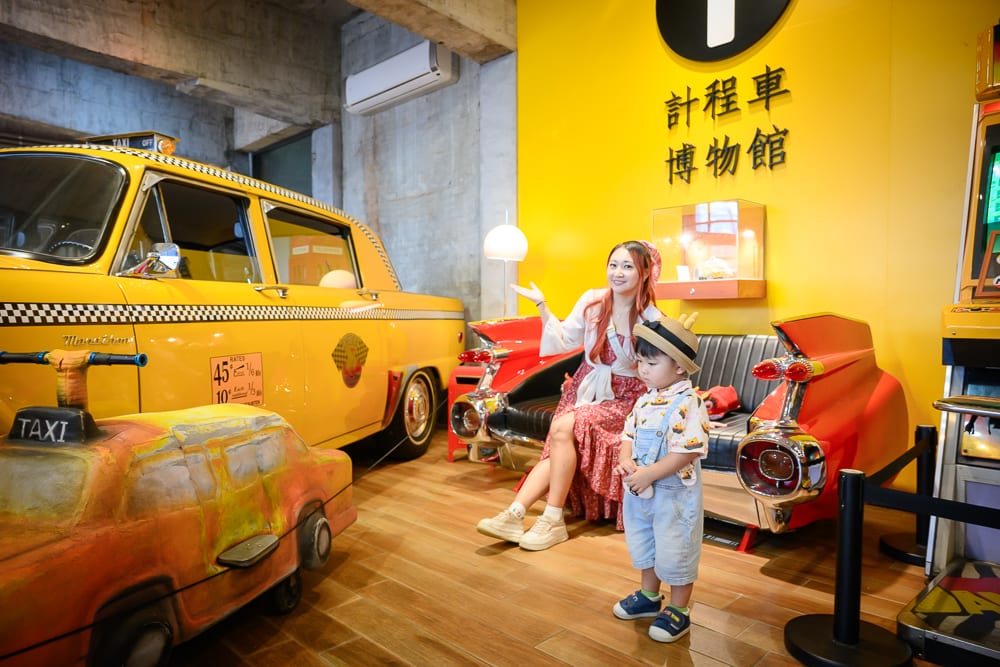 宜蘭計程車博物館｜室內、計程車博物館、黃牆、古董車、沙發、女人、小孩、木地板。女子與幼童坐在由車尾改裝的黑色沙發上，前方停放黃色古董計程車與小型玩具車，牆面懸掛「計程車博物館」黑色字樣。