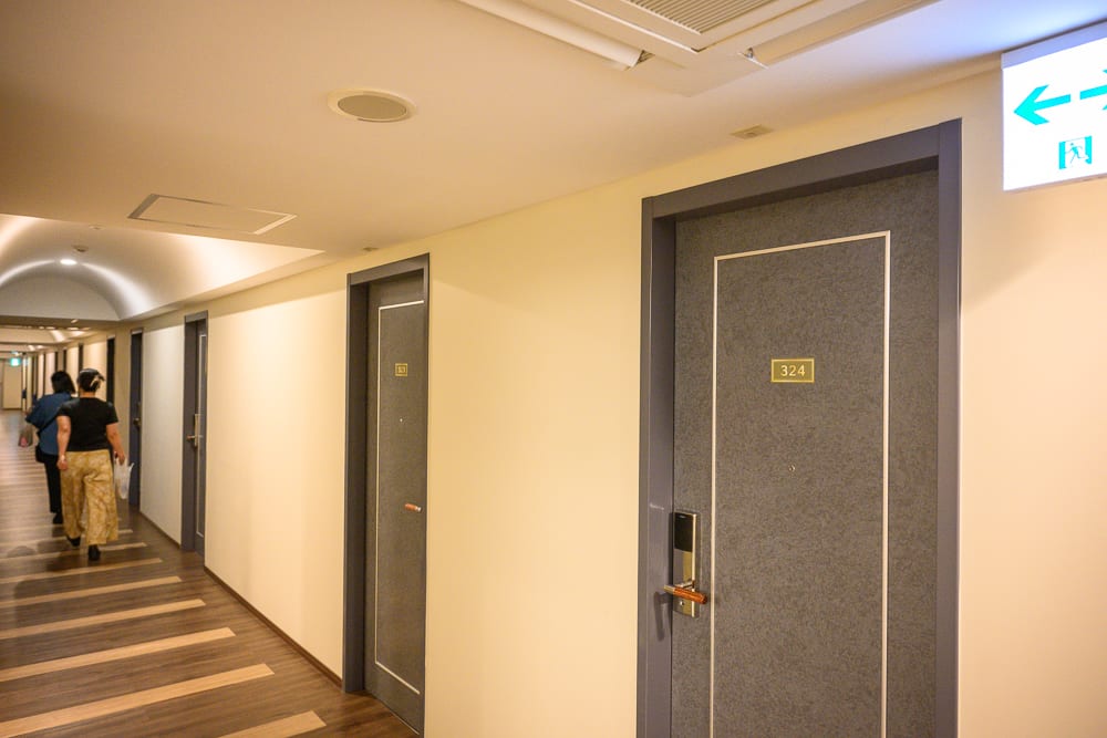 日本沖繩喜璃愈志LCH渡假村|建築外觀、戶外泳池與周邊補給 10 日本沖繩喜璃愈志LCH渡假村|室內、走廊、房門、木地板,米色牆面嵌著深灰色門框。兩名行人在長廊深處背對鏡頭前行,天花板裝設圓形崁燈與白色消防指示牌,映出冷色調的人工照明。