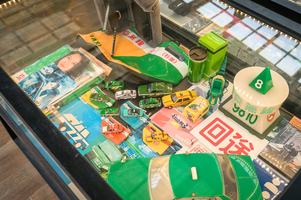 宜蘭計程車博物館｜玻璃展示櫃內，玩具車模型、雜誌、綠色保溫杯、迴轉壽司風格的數字指示牌與車頂燈罩等物品鋪滿檯面， overhead 燈光照射著。