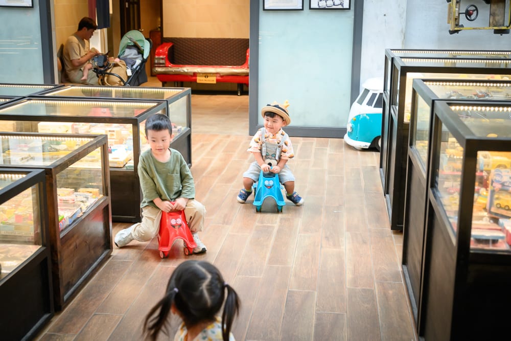 宜蘭計程車博物館｜室內，孩童，玩具車，展示櫃，木地板。兩名幼童分別騎乘紅色與藍色滑步車，行駛於陳列玻璃櫃間的木紋地板走道，背景擺放紅色復古車椅與藍白小型車模型。