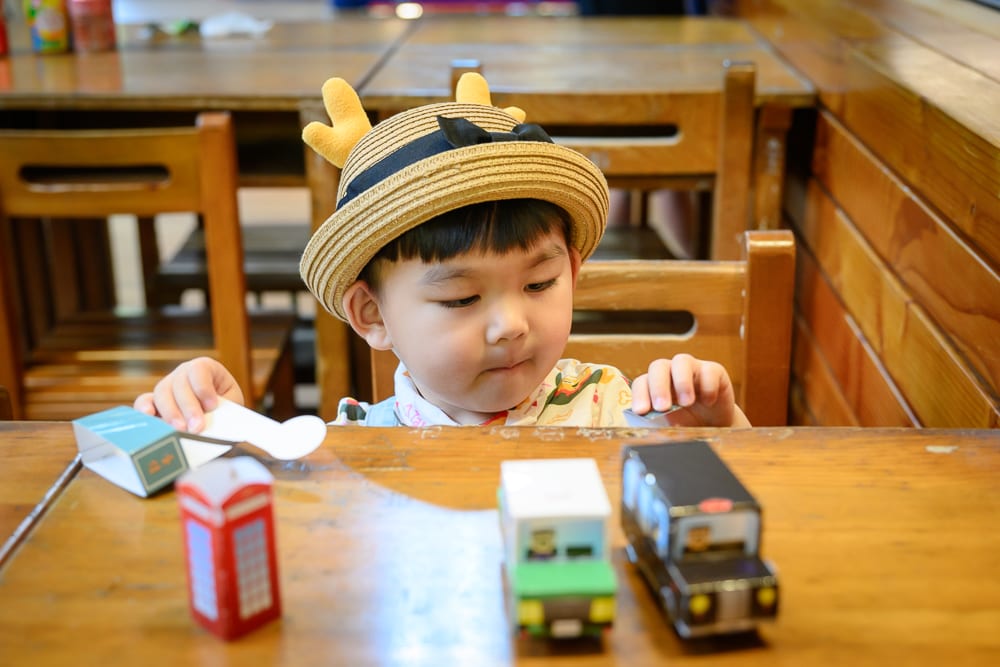 宜蘭計程車博物館｜小孩、紙模型、電話亭、木桌、室內，戴著鹿角草帽的幼童坐在木製桌椅前，專注組裝桌上的紙模型，周圍擺放小型車輛與電話亭紙雕。
