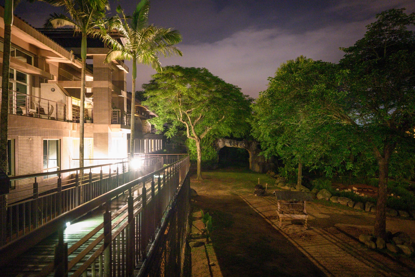 新竹六福莊關西｜夜間建築外觀，金屬欄杆走廊，棕櫚樹，茂密綠樹，草地小徑，石洞入口，戶外燈光照明。