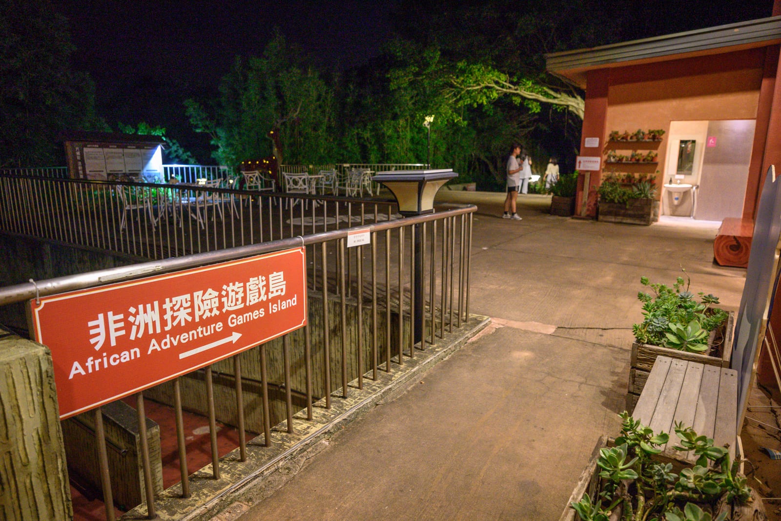 新竹六福莊關西｜夜間戶外露臺，鐵欄杆旁掛著紅底白字的「非洲探險遊戲島」指示牌，階梯通往地下。右側為建物入口，擺放植栽木箱與長凳，背景有茂密樹木與遠方遊客。