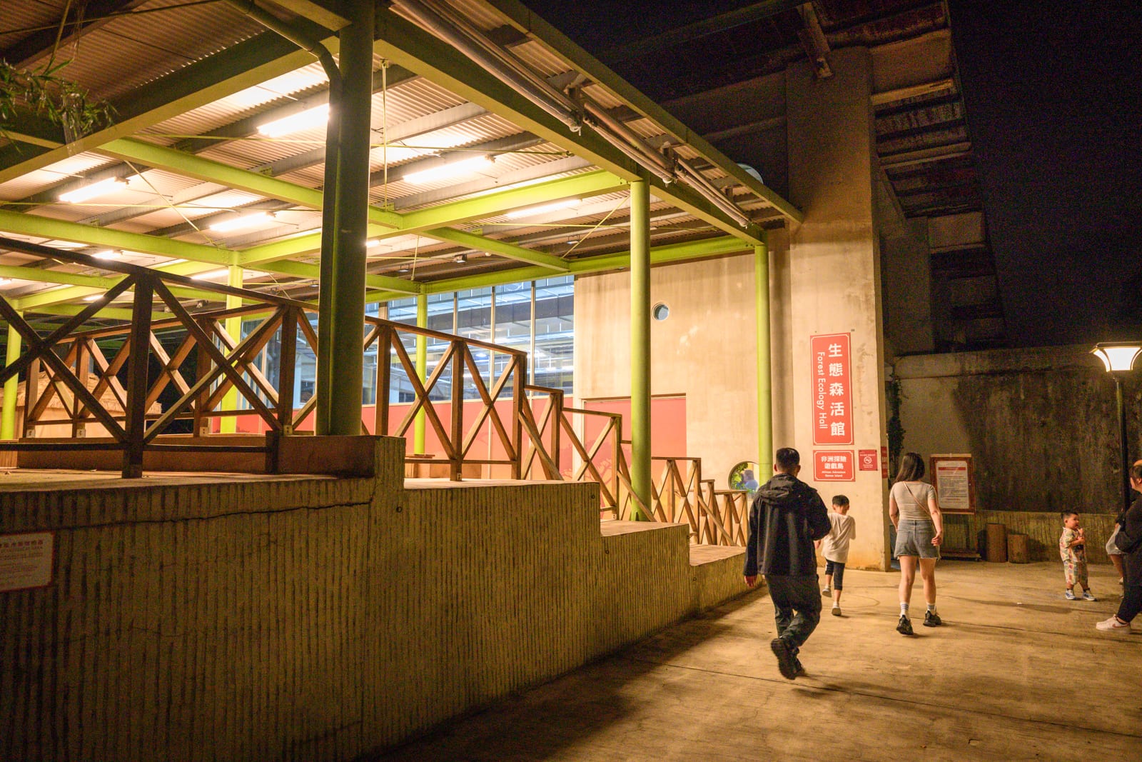 新竹六福莊關西｜夜間建築外側走廊，綠色鋼構屋頂下方安裝多管日光燈，混凝土階梯與木質欄杆。幾位行人穿梭於水泥地坪廣場，右側牆面懸掛紅底白字指標，遠處立著一盞路燈。
