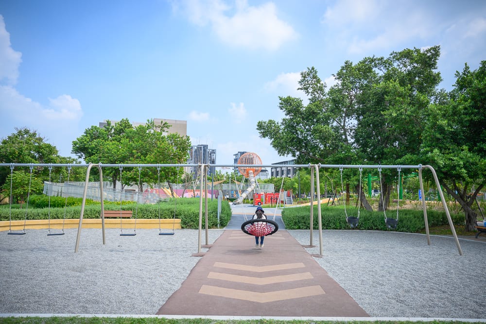 彰化員林兔馬鹿公園完整指南|親子遊戲場玩法解析 2 彰化兔馬鹿公園|戶外公園,遊樂場,盪鞦韆,網狀圓形鞦韆,長椅,綠樹,草地,藍天白雲,遠方建築。一名孩童正坐在金屬支架的圓形網狀鞦韆上盪著,地面鋪滿細小碎石,周圍有綠色植栽與成排樹木。