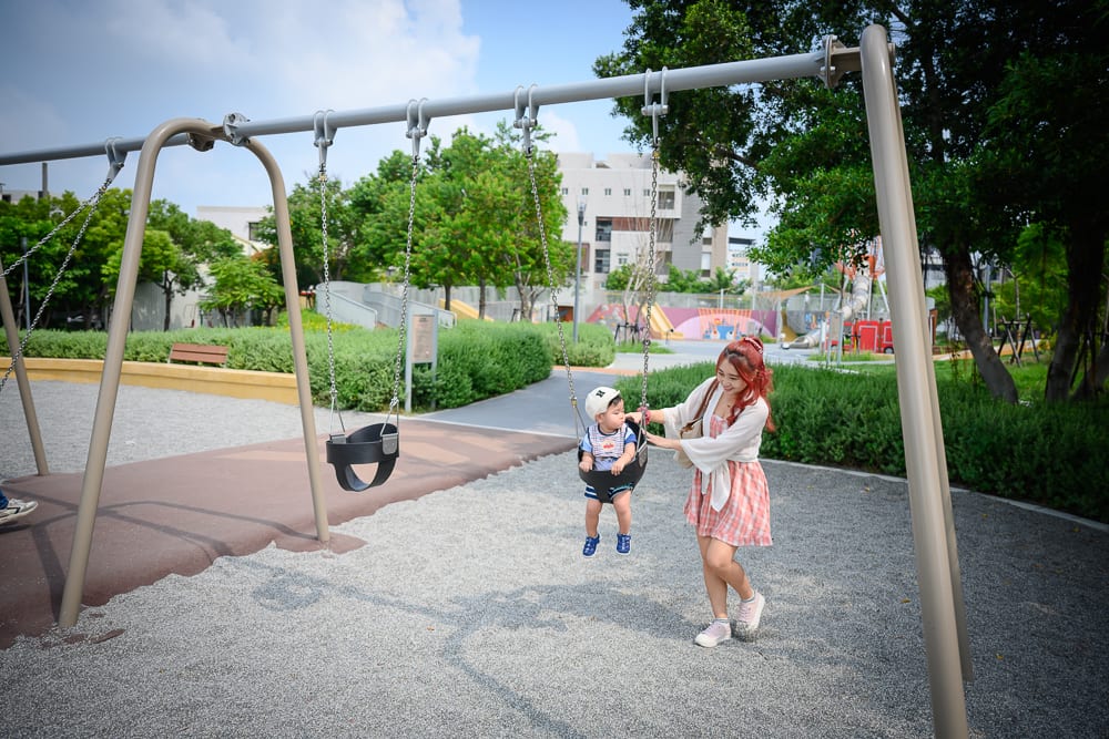 彰化員林兔馬鹿公園完整指南|親子遊戲場玩法解析 13 彰化兔馬鹿公園|金屬盪鞦韆、公園、綠樹、碎石地、女子、男童,女子站在盪鞦韆旁,伸手輕觸坐在座椅內的男童,背景有草地與建築物。