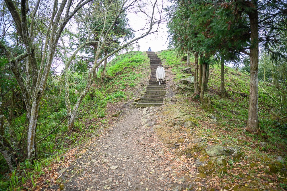 嘉義寒溪呢｜泥土小徑，階梯，坡地，林木，穿白色雨衣的人，綠草。背對鏡頭的人正向上走，兩側佈滿樹木與枝葉，灰白天空
