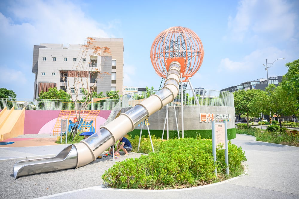 彰化員林兔馬鹿公園完整指南|親子遊戲場玩法解析 7 彰化兔馬鹿公園|戶外公園,大型遊具,溜滑梯,球形攀爬架,建築物,草皮,綠樹,步道,藍天白雲。透明管狀溜滑梯延伸至沙坑地面,周圍環繞灌木叢與低矮植栽,遠方有多層樓房與路燈。