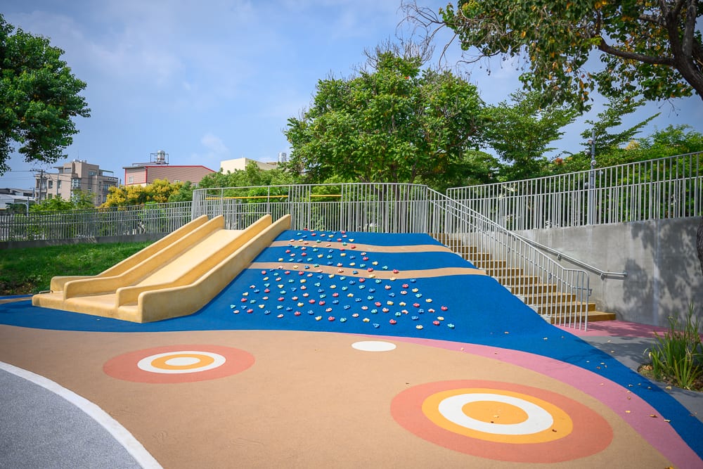 彰化員林兔馬鹿公園完整指南|親子遊戲場玩法解析 6 彰化兔馬鹿公園|戶外公園,溜滑梯,攀岩牆,階梯,金屬欄杆,安全地墊,綠樹,藍天。色彩鮮明的兒童遊樂設施設置於坡面上,周圍環繞灰色水泥矮牆與欄杆,地面鋪設帶有同心圓圖案的防跌落軟墊,上方枝葉茂密。