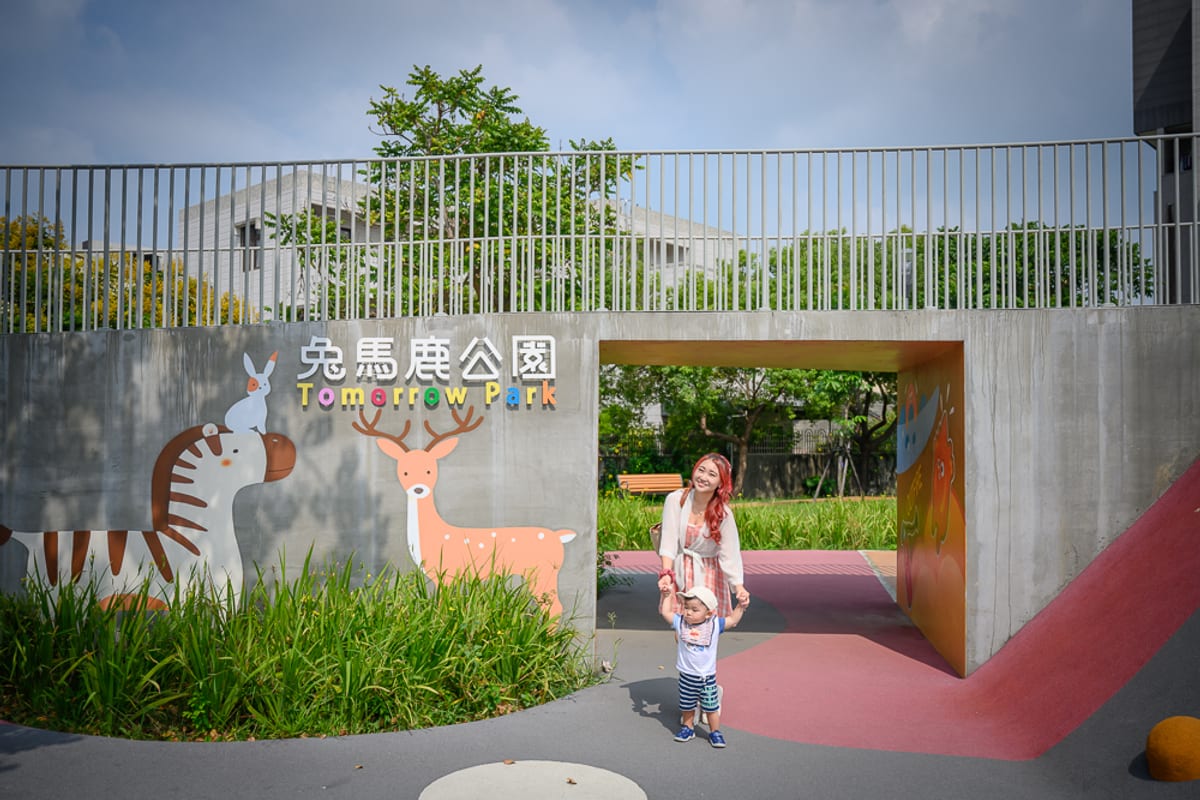 彰化員林兔馬鹿公園完整指南|親子遊戲場玩法解析 1 彰化兔馬鹿公園|女性與小孩、公園、灰牆、動物壁畫、欄杆、綠樹、通道,灰色水泥地面與紅磚道相接,立體招牌顯示文字「兔馬鹿公園 Tomorrow Park」。